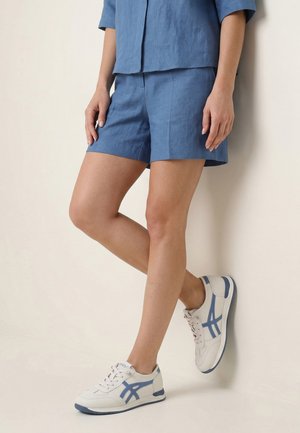 Osoba nosząca niebieskie szorty i pasującą koszulę z białymi sportowymi butami z niebieskimi akcentami, stojąca na neutralnym tle.