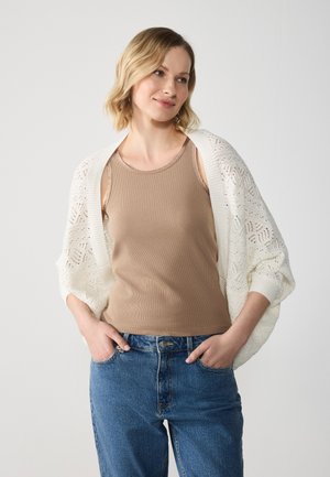 Langlopende, open-gehaakte cardigan in het wit over een lichtbruine tanktop; blauwe hoog-waisted jeans met een relaxte pasvorm.
