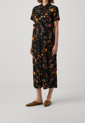 Femme portant une robe midi noire avec un motif floral coloré, manches courtes, col, ceinture à la taille, et chaussures plates marron à bouts pointus sans lacets.