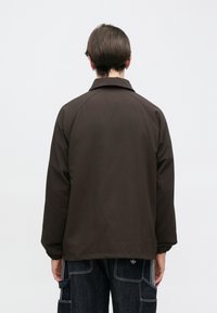 Καφέ μπουφάν με κλασικό γιακά και ελαστικά μανίκια, κατασκευασμένο από λείο ύφασμα. Προβολή από πίσω, με χαλαρή εφαρμογή και χωρίς ορατά μεταλλικά στοιχεία.