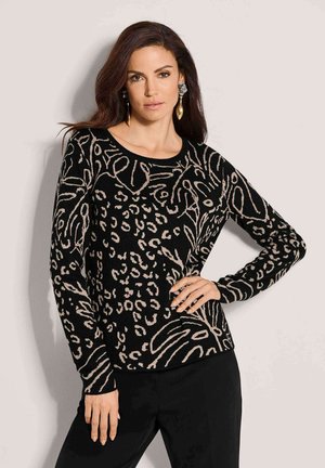 Femme aux longs cheveux foncés portant un haut à manches longues noir et beige à motifs et un pantalon noir, posant avec une main sur la hanche.