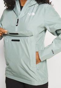 Light teal waterproof jacket with a hood, zippered front pocket, and adjustable cuffs. Features a logo on the left chest and side pockets.