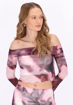 Ung kvinde med langt, bølget hår iført pink og lilla tie-dye crop top med bare skuldre og lange ærmer samt matchende bukser, der ser til siden.