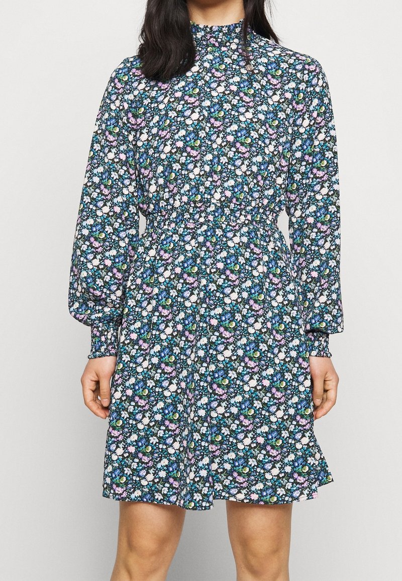 Femme portant une robe à manches longues, longueur genou, avec un col montant et un motif floral coloré dans les tons bleu, blanc et violet.