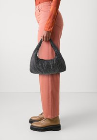 Sac à épaule à pois noir et blanc avec une forme douce et arrondie, en tissu texturé, tenu par une personne portant un pantalon et des chaussures roses.