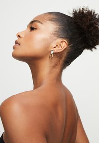 Pendientes en tono dorado con una piedra blanca triangular y un acento circular, sobre un tono de piel suave en un perfil lateral.