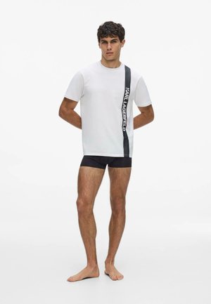 Muž stojí bosý, oblečený v bielom tričku s vertikálnym čiernym prúžkom a nápisom „KARL LAGERFELD“, v kombinácii s čiernymi šortkami, na bielom pozadí.