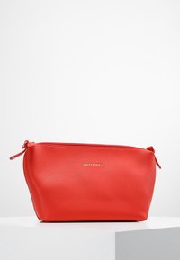 Punainen mukulakivinen nahkapussi, jossa on kultainen vetoketju ja pieni kahva. Edessä on kohokuvioitu logo. Sileä pinta, soikea muoto.