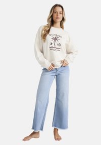 Sudadera crema con gráfico de palmera y texto, combinada con jeans de pierna ancha azul claro con dobladillo deshilachado. Modelo posando descalzo.