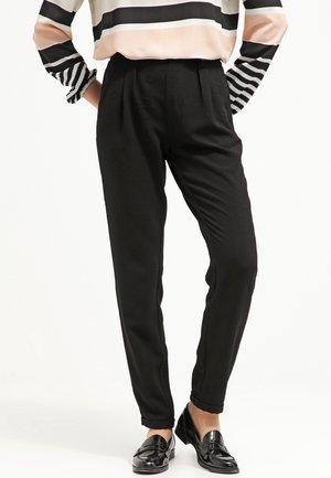 Sorte skræddersyede bukser med ankellængde, båret med en langærmet top med striber og sorte loafers, hænderne hvilende på hofterne.