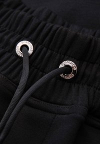 Schwarzer elastischer Bund mit schwarzem Kordelzug, der durch silberne Metallösen mit der Gravur "OMBREWEAR" gezogen ist.