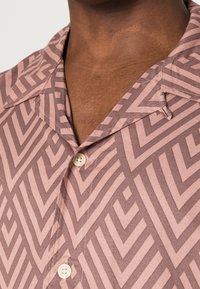 Närbild av brun hud som bär en skjorta med krage i rosa och mauve chevronmönster, med vita knappar fastknäppta vid bröstet.