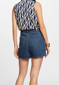 Mujer vistiendo una blusa sin mangas con estampado y pantalones cortos de mezclilla azul de tiro alto, con una pulsera dorada en su muñeca derecha, de espaldas.