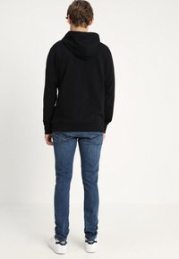 Felpa nera con cappuccio, dal fit rilassato, abbinata a jeans blu slim e sneakers bianche. La texture sembra morbida e informale.