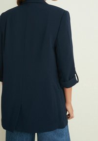 Blazer entallado de color azul marino con mangas de tres cuartos, puños enrollados y una abertura lateral. Fabricado con una tela suave y con un diseño minimalista.