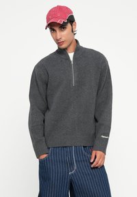 Grijze zip-up trui met een geribbelde onderrand en mouwen, gemaakt van een gestructureerde stof. Gecombineerd met blauw gestreepte broek en een roze baseballpet.