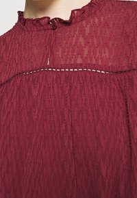 Burgunderfarbene Bluse aus einem strukturierten, durchsichtigen Stoff. Verfügt über einen gerüschten Kragen, eine Knopfleiste und horizontale Stickakzente entlang der Naht.