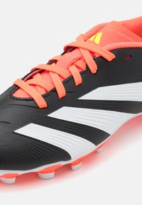 Botín de fútbol negro con una textura suave, que presenta cordones de color naranja brillante, una lengüeta de malla y rayas blancas en el costado. Clavos de goma en la suela.