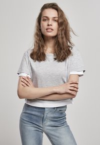 Harmaa lyhythihainen cropped-t-paita valkoisilla yksityiskohdilla, valmistettu pehmeästä kankaasta. Yhdistettynä vaaleansinihin farkkuihin, mallilla on kädet ristissä.