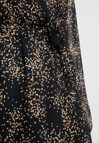 Svart klänning i genomskinligt texturerat tyg med ett spritt mönster av beige prickar. Knutit midja-detaljen framhäver formen.