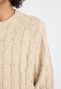 Pulover s kabelskim pletenjem v bež barvi z večbarvnimi pikicami. Ima okroglo ovratnik in teksturiran dizajn, ki prikazuje udoben pleteni videz.