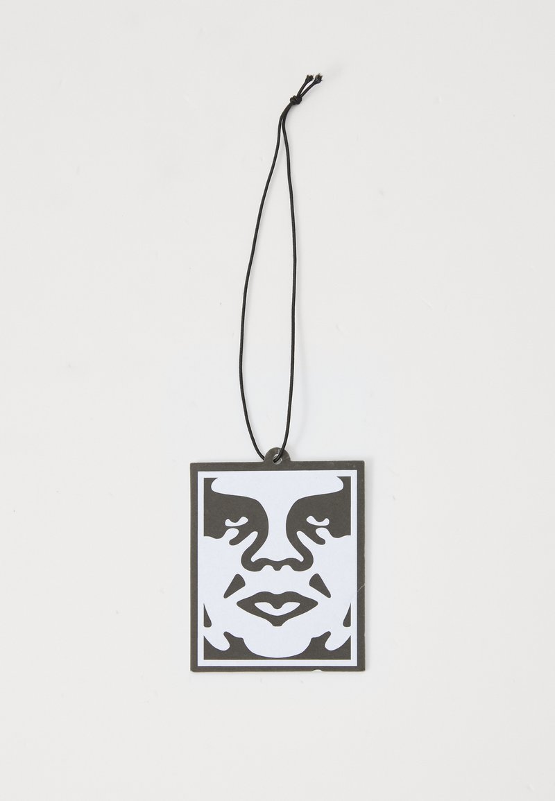 Etiqueta gráfica em preto e branco com um design de rosto estilizado, forma quadrada, com um cordão preto fino para pendurar. Acabamento mate, textura mínima.