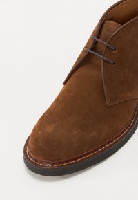 Bruine suède enkelboot met een ronde neus, dubbele bruine veters en een gelaagde zool met een contrasterende stikselafwerking.