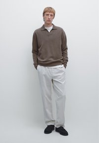 Brown knitted sweater with a collared neck, paired with light beige trousers and black sneakers. Minimalist background.