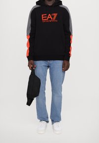 Sudadera con capucha negra con acentos en gris y naranja, que presenta un logo de EA7. Combinada con vaqueros azul claro y zapatillas blancas. Sosteniendo una bolsa negra.