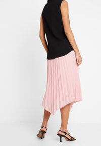 Musta hihaton paita, jossa on korkea kaulus, yhdistettynä vaaleanpunaiseen laskostettuun hameeseen, jossa on kaareva helma ja mustat remmipohjalliset kengät.