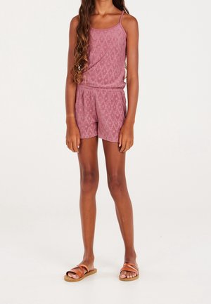 Jeune personne portant une combinaison-short sans manches mauve à motifs et des sandales marron à lanières, debout devant un fond uni.