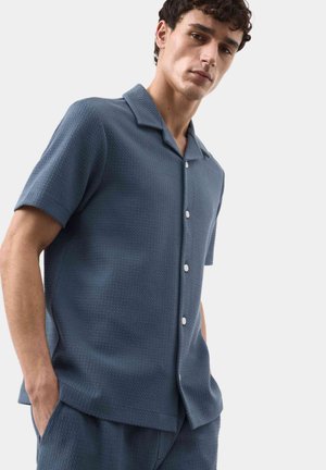 Joven con camisa azul de manga corta con textura y botones, y pantalones a juego, con las manos en los bolsillos, frente a un fondo liso.