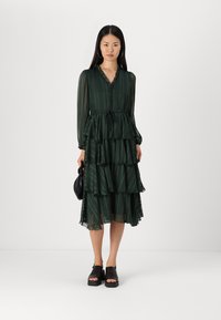 Mörkgrön, lager-på-lager klänning med knäppning framtill, långa ärmar och ett bälte i midjan. Semi-transparent tyg med subtila ränder. Svarta skor.