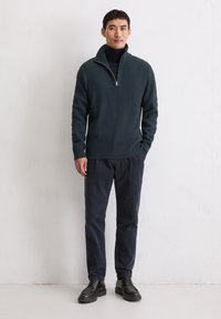 Donkergroen geribbeld sweater met een ritssluiting kraag, gecombineerd met marineblauwe corduroy broek en zwarte veterschoenen. Soepele texturen en gestructureerd ontwerp.