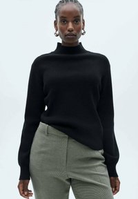 Maglione nero a maglia con collo alto e maniche lunghe, abbinato a pantaloni verdi chiari testurizzati. Presenta una vestibilità rilassata.