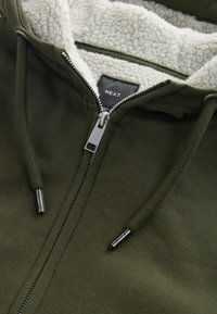 Ritsvest in olijfgroen met een zachte, crèmekleurige fleece voering. Voorzien van een zwart label met de tekst "NEXT" en zilverkleurige ritssluiting.