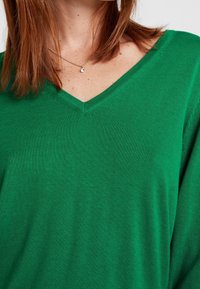 Grüner V-Ausschnitt-Pullover aus weichem Textil; verfügt über eine dezente Textur und eine lockere Passform mit mittellangen Ärmeln. Schlichtes Design ohne Muster.