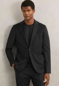 Sort skræddersyet blazer med notch-lapel, enkelt knaplukning og to frontlommer. Båret over en sort t-shirt med rund halsudskæring. Glat tekstur.