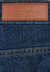 Denim tkanina tamno plave boje, koja sadrži smeši kožni znak s utisnutim natpisom "URBAN CLASSICS DENIM FACTORY" i kontrastnim šavovima.