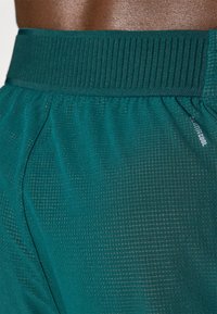 Shorts de sport teal avec une taille côtelée et un tissu texturé présentant un motif à léger quadrillage. Comprend le branding "UltraCool".