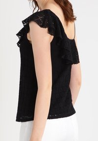 Svart spetsöverdel med volangdetaljer på axlarna och en strukturerad mönster, med en figursydd silhuett och tunna justerbara axelband.