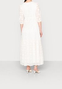 Abito maxi bianco con dettagli in pizzo intricati, maniche a tre quarti, gonna a strati e una texture liscia. Abbinato a tacchi di colore chiaro.