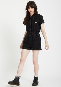 Mono de mezclilla negra con mangas cortas, cinturón en la cintura y textura estampada. Presenta un logo en el pecho. Se lleva con botas negras de cordones.