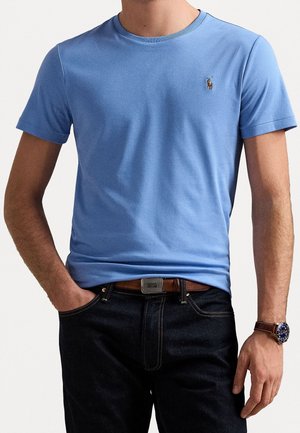 Mężczyzna w jasnoniebieskim t-shircie z krótkim rękawem z małym wyszywanym logo, ciemnych jeansach, brązowym pasku i zegarku na rękę z brązowym skórzanym paskiem.