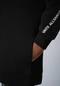 Sweatshirt noir en mélange de coton lisse avec poignets et ourlet côtelés. Comprend une poche et un texte imprimé blanc le long de la manche.