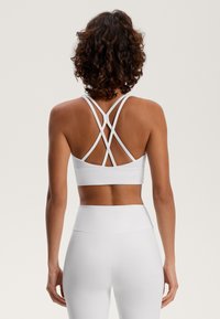 Biela športová podprsenka s prekríženými ramienkami na chrbte, vyrobená z pružného materiálu bezšvového dizajnu s jemným logom na spodnej strane.