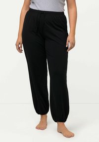 Schwarze Hose mit elastischem Bund und Kordelzug, eng zulaufenden Säumen und lockerer Passform, aus weichem Material. Barfuß getragen mit einem grauen Oberteil.