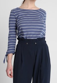 Mörkblå, högmidjade byxor med veck och knapppynt; kombinerat med en långärmad topp i blå ränder som har en båturringning.