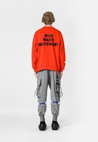 Sudadera roja con texto negro "MOD WAVE MOVEMENT" en la parte posterior; combinada con pantalones cargo grises que presentan bolsillos y detalles de parches. Calzado negro.