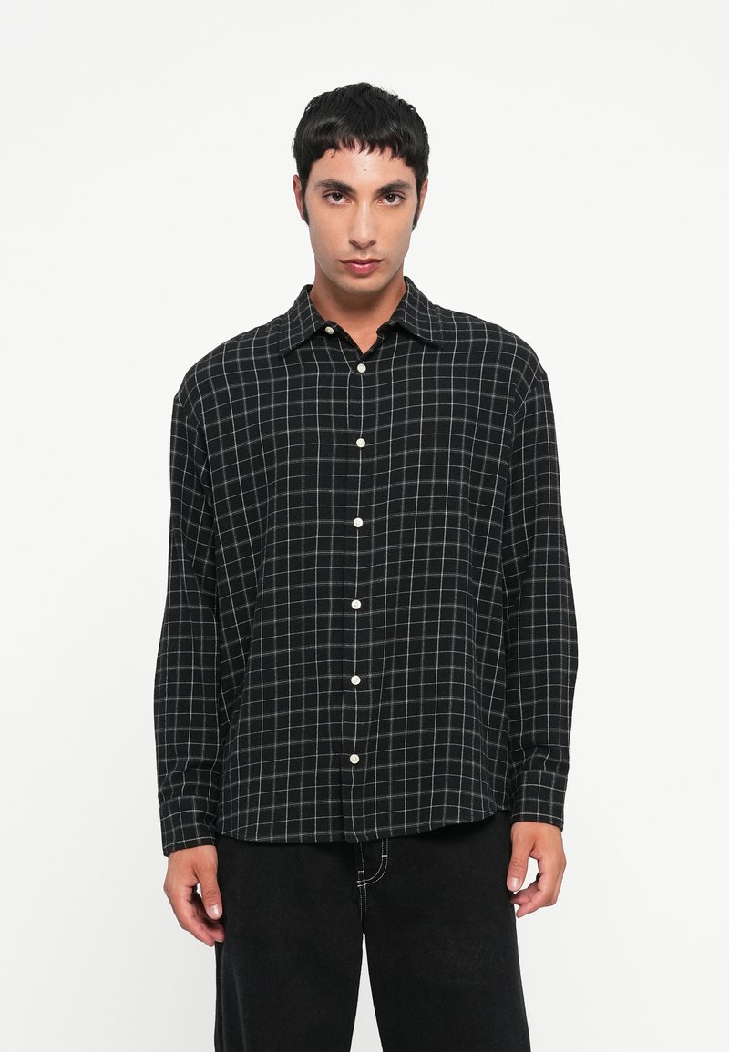 Czarna i biała koszula w kratę z rozłożystym kołnierzykiem, długimi rękawami i białymi guzikami. Noszona z ciemnymi spodniami, miękka faktura materiału.